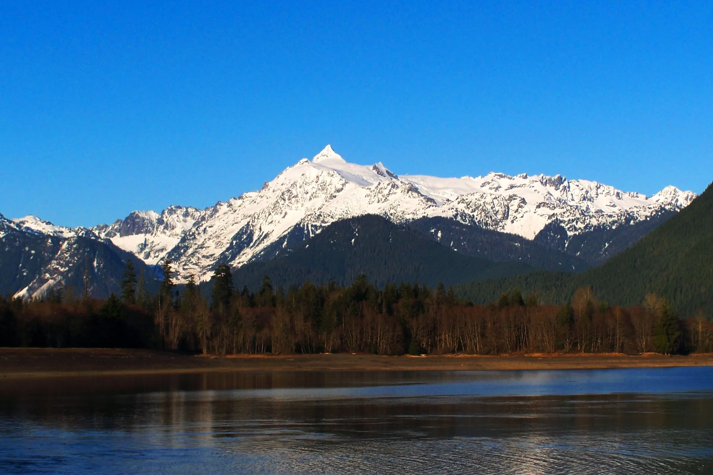 Snow-covered peak above forested ridges reflected in the still water of Baker Lake in winter